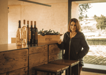 Foto da Ana Patrícia em uma vinícola com uma taça de vinho em mãos e garrafas na mesa