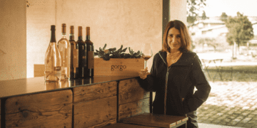 Foto da Ana Patrícia em uma vinícola com uma taça de vinho em mãos e garrafas na mesa